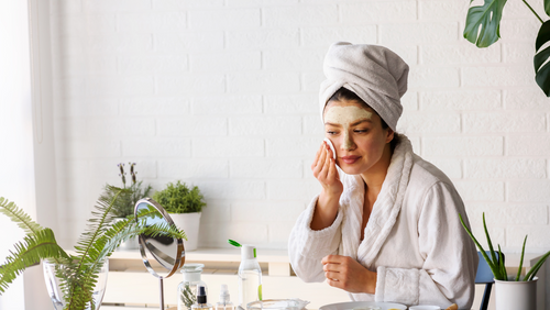 Woman applying facial mask with towel on head in a bright room with plants